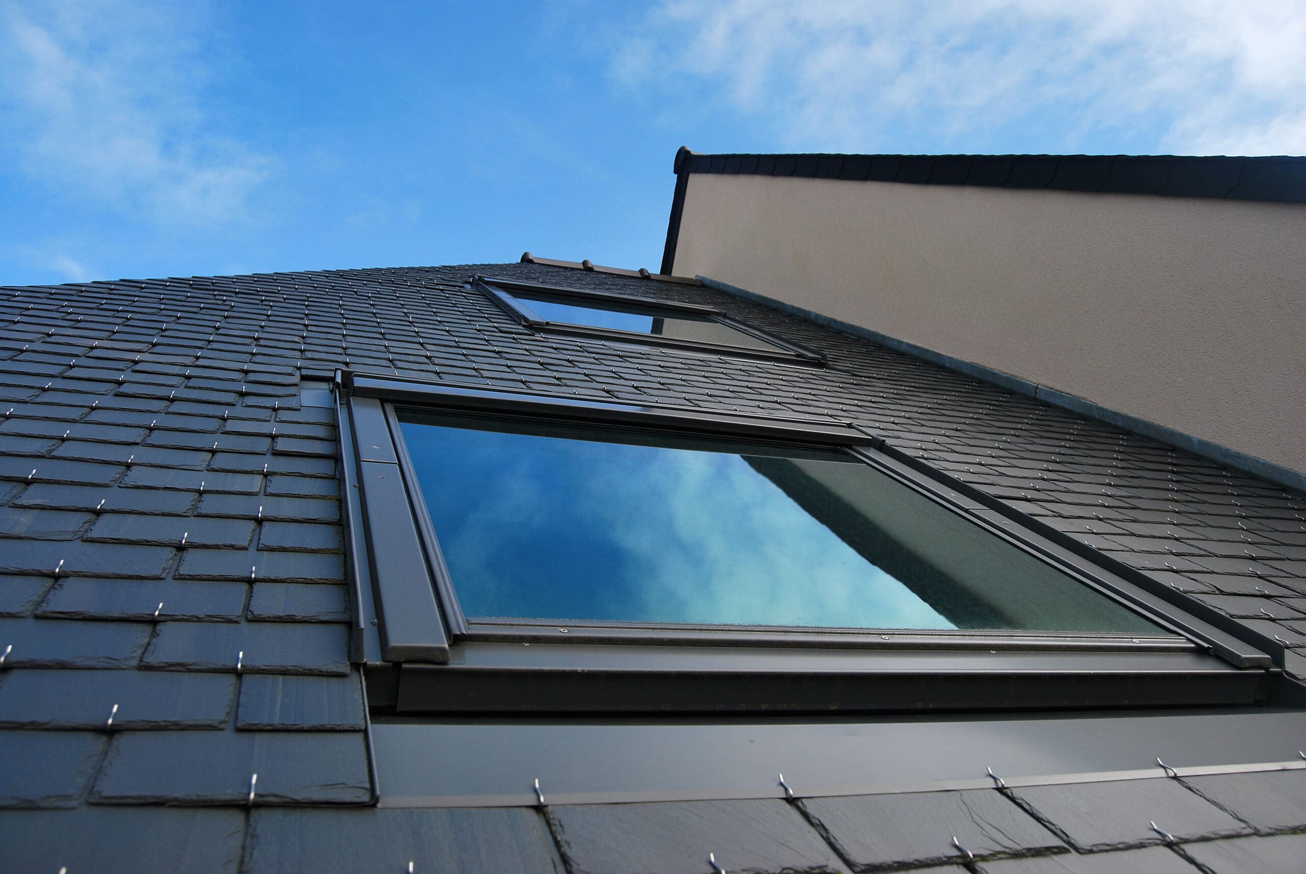 A low-angle view of a modern building with a slate tiled roof and two large windows reflecting a partly cloudy blue sky. The building has beige walls and sharp angles, creating a sleek architectural design.