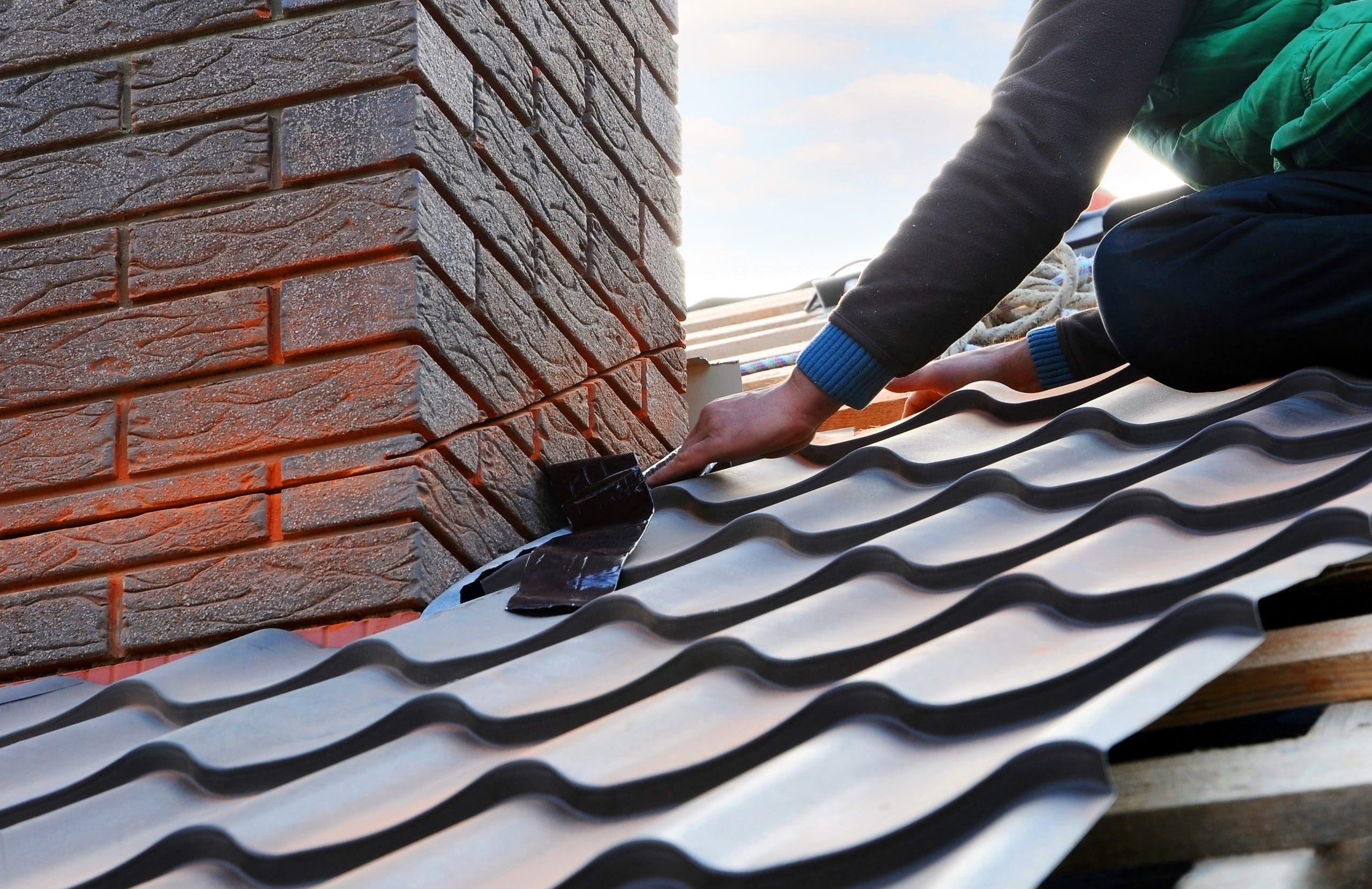 A person working on a metal roof, fitting it around a brick chimney. They are using tools and adjusting the roofing material. The roof features a corrugated pattern, and the setting appears to be outdoors under a clear sky.