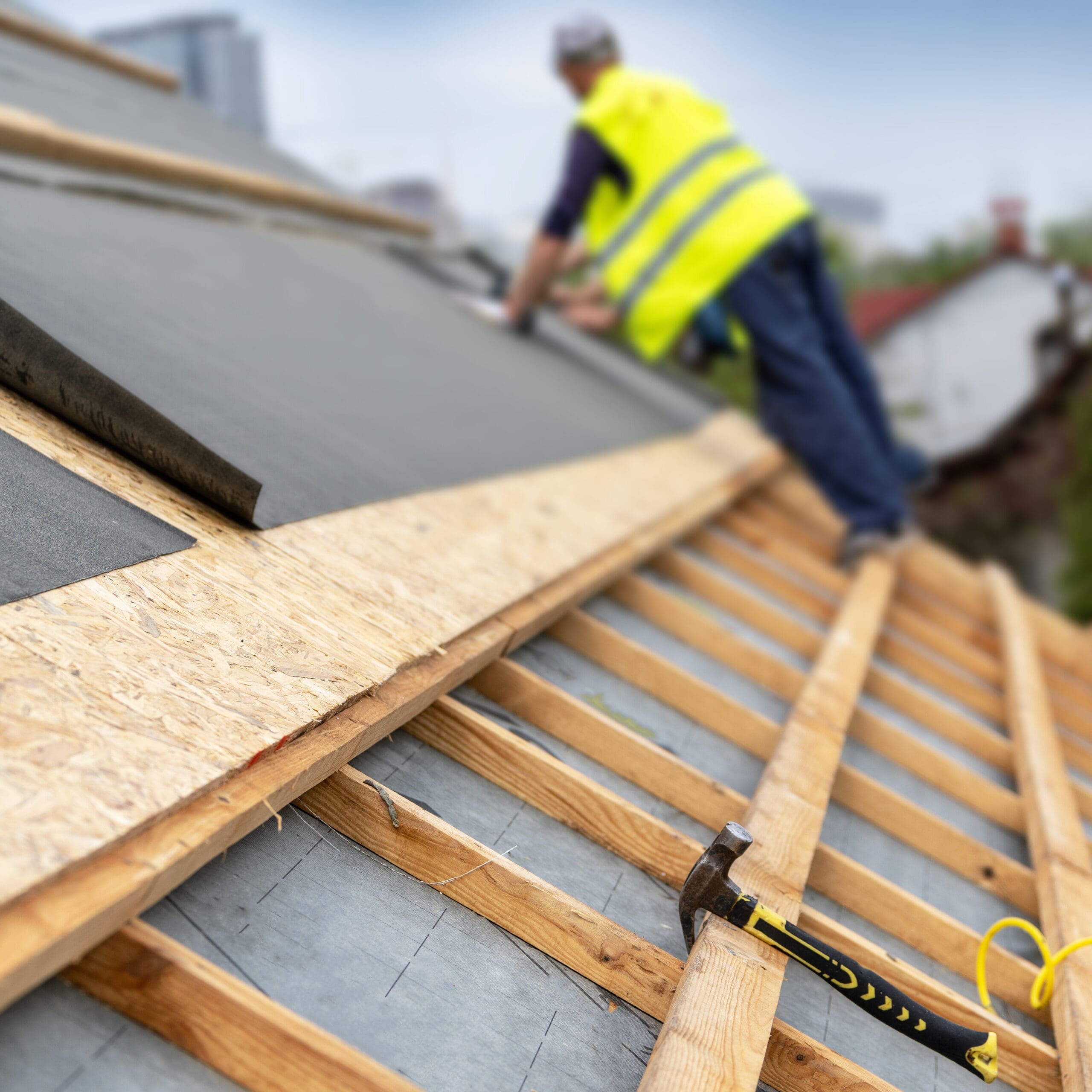 A construction worker wearing a neon yellow safety vest is working on a slanted rooftop. Wooden beams and roofing materials are visible, along with a hammer resting on the beams. The background shows a blurred urban landscape.