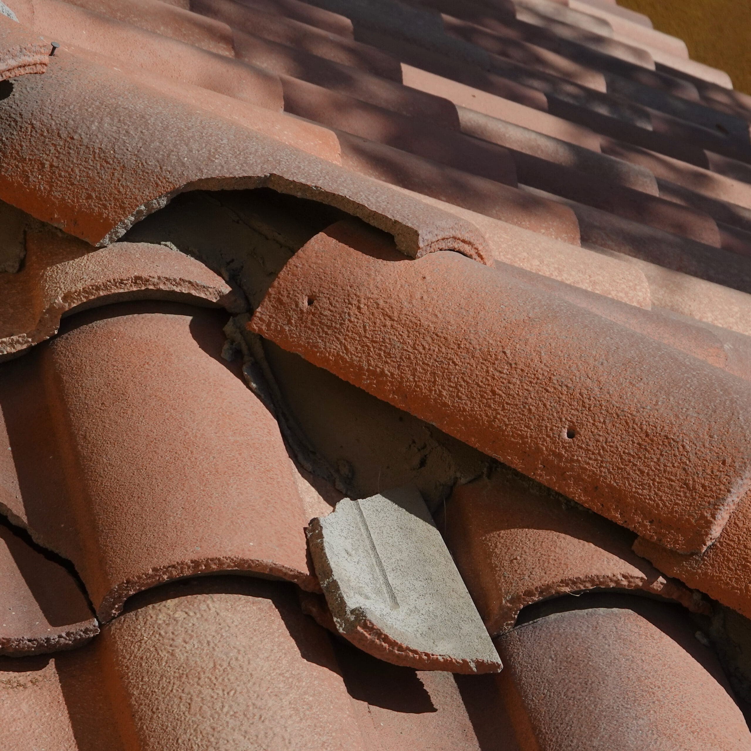 Close-up of a damaged terracotta roof with broken tiles. Several tiles are cracked and misaligned, exposing gaps. The surrounding roof appears sunlit, casting shadows between the rows of tiles.