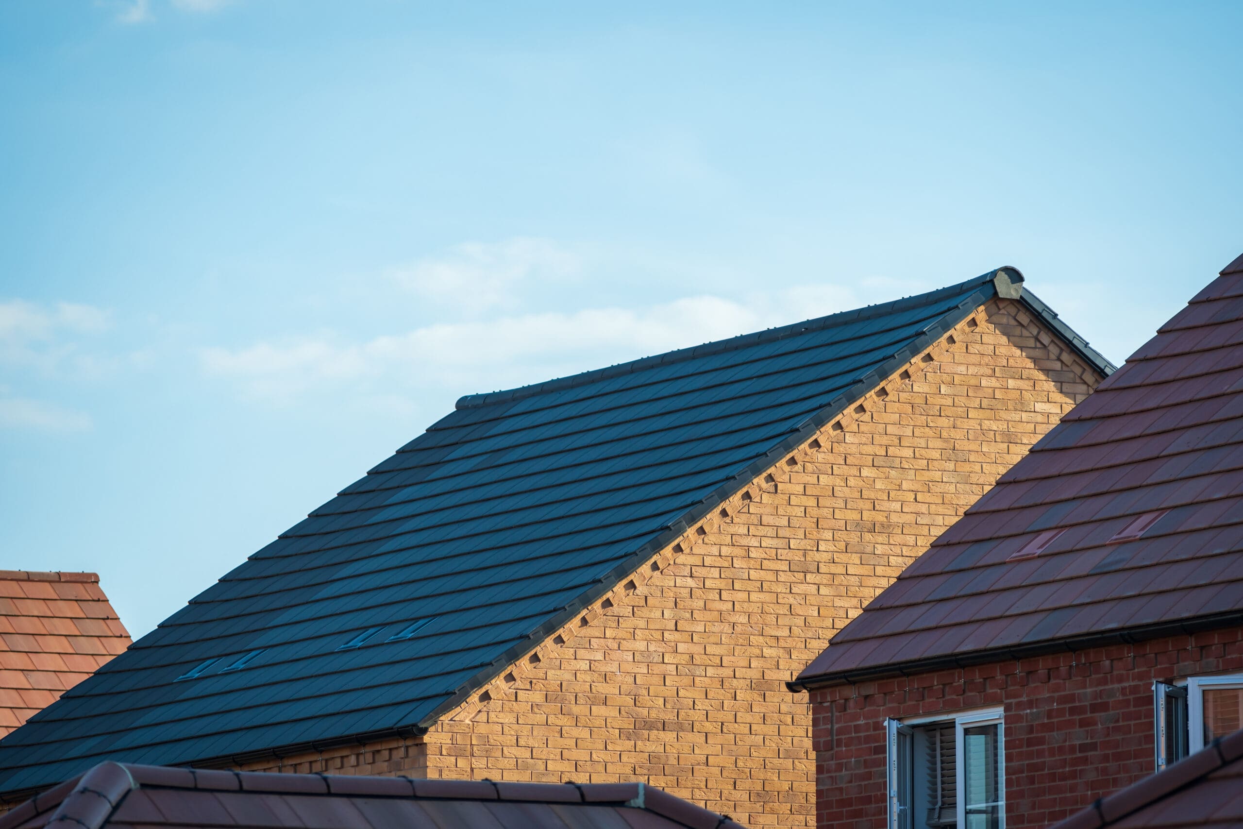 A view of several house rooftops, primarily featuring a brick house with a gable roof. The roofs are in shades of dark and reddish-brown. The background is a clear blue sky with a few light clouds.