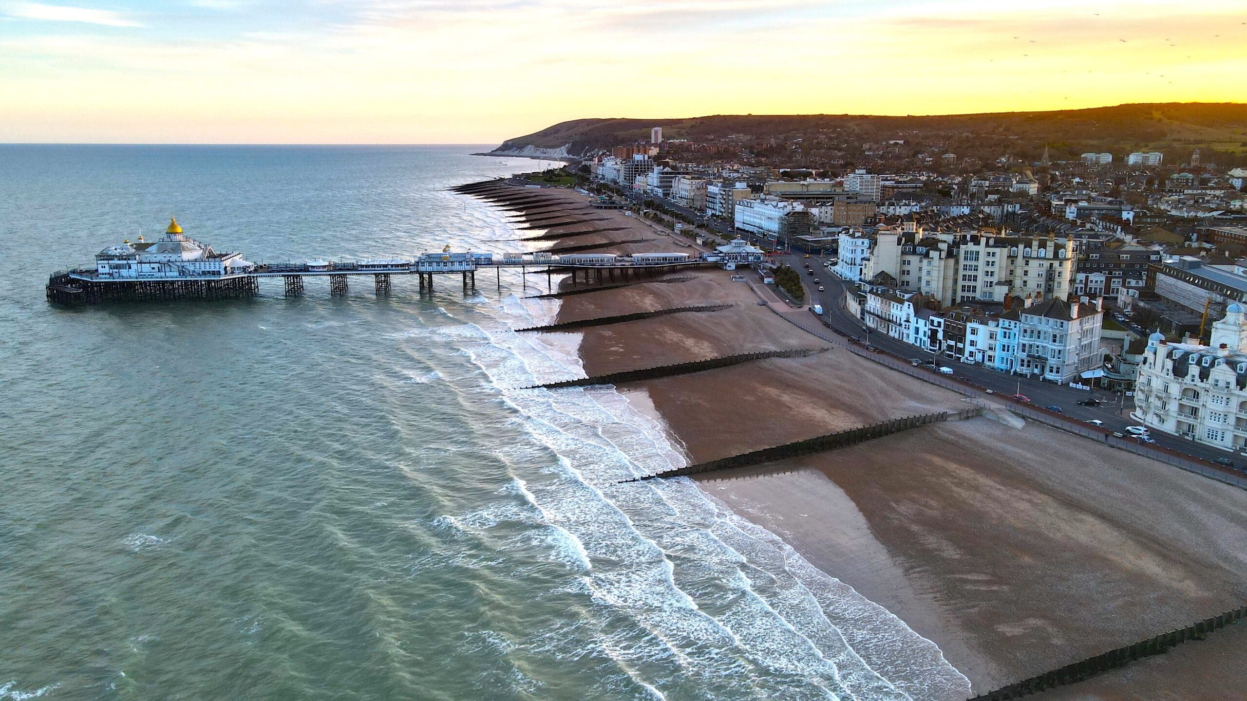 A coastal town at sunset with a pier extending over the ocean. Waves gently lap the sandy beach lined with wooden groynes. Buildings and roads parallel the shoreline, with rolling hills in the background under a colorful sky.