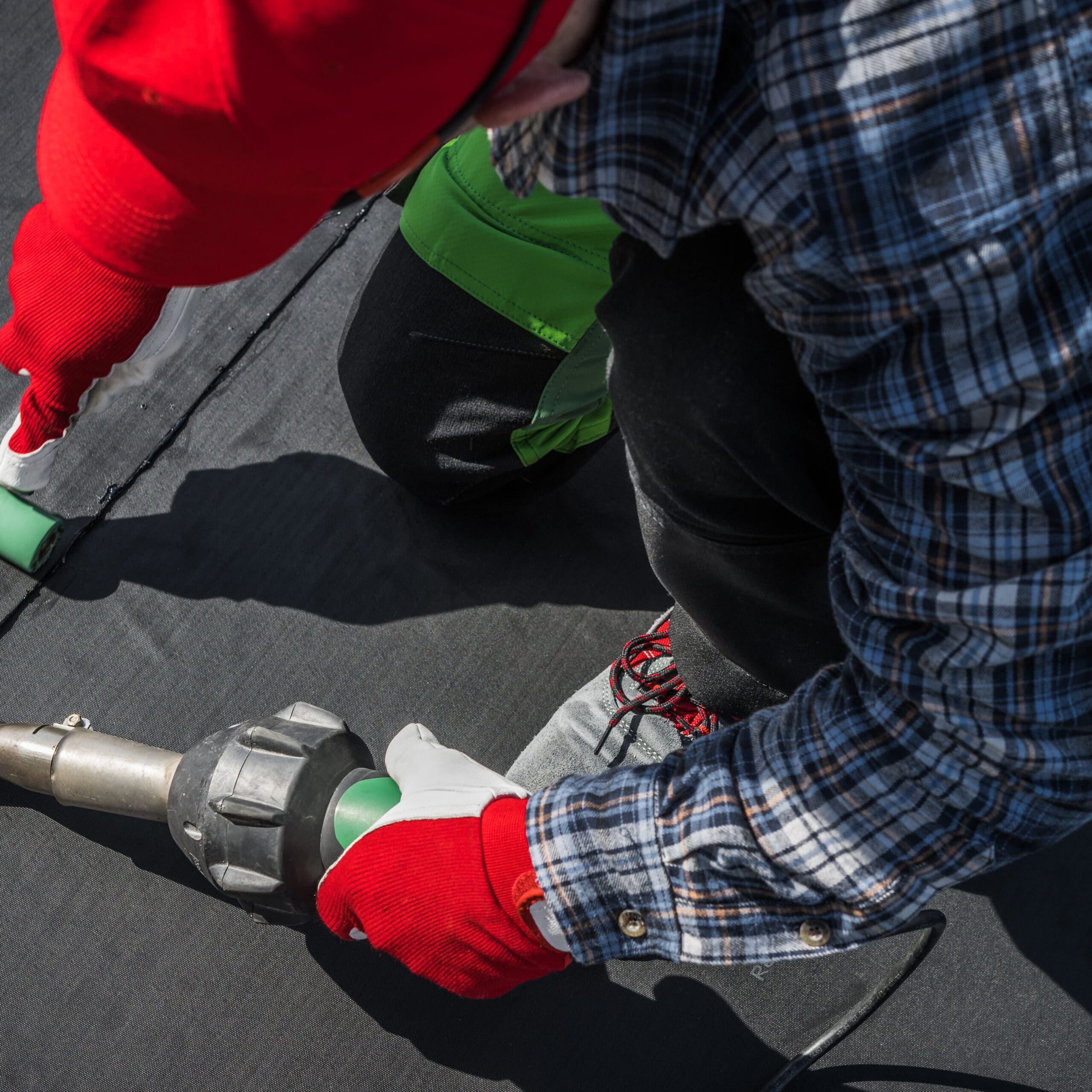 A person wearing a red hat and gloves is using a tool to work on a dark surface, possibly sealing or welding. They are dressed in a plaid shirt and green pants, and the scene is focused on their hands and the tool.