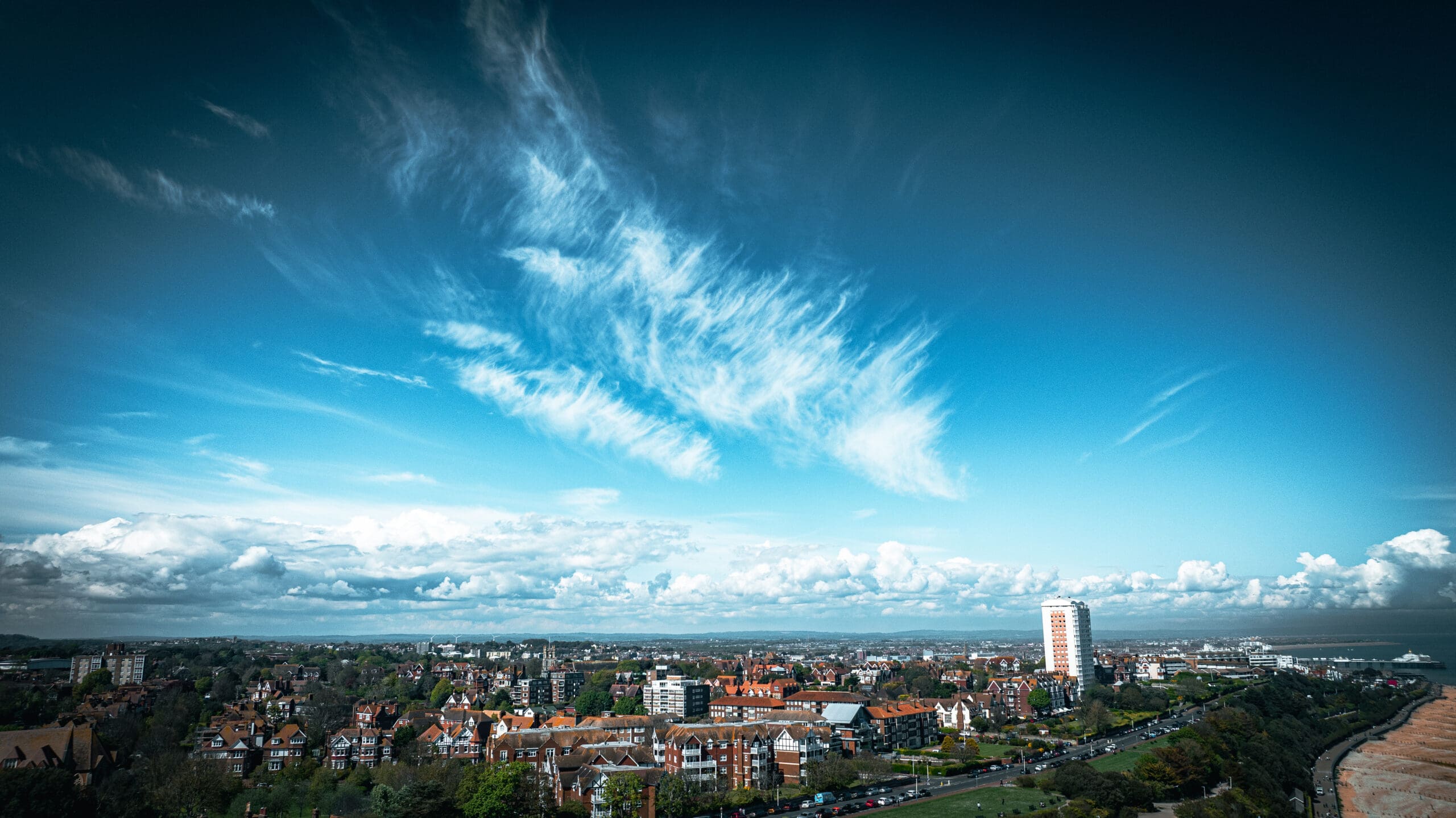 Aerial view of a cityscape with a prominent tall building and rows of houses. The sky is bright blue with scattered, wispy clouds. Green fields and a coastline are visible in the foreground.