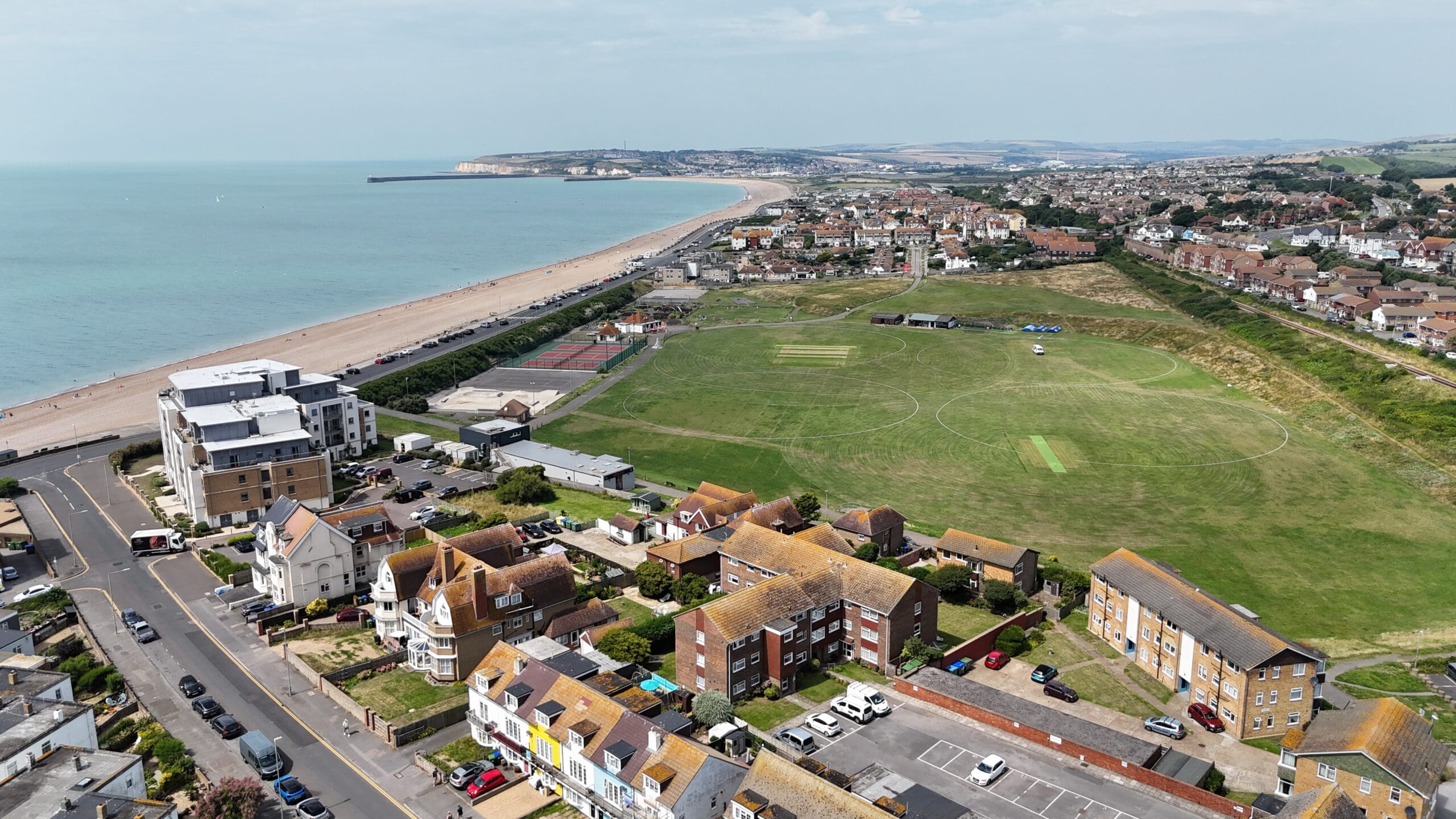 Aerial view of a coastal town with a large grassy cricket field in the center. Surrounding buildings include houses and apartments with brown roofs. The beach and ocean are visible in the background under a cloudy sky.