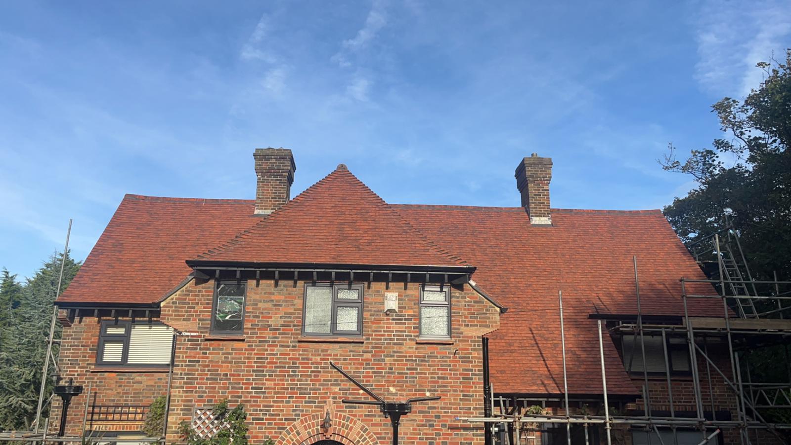 A two-story red brick house with a steep red-tiled roof and two chimneys. Scaffolding is partially set up around the building, and the sky is clear and blue.