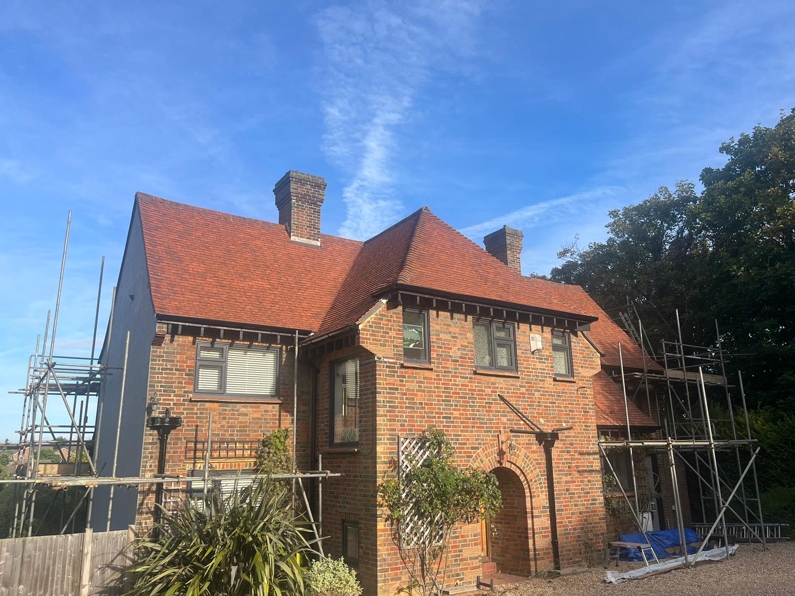 A two-story brick house with a red-tiled roof, featuring an arched entryway and two chimneys. Scaffolding is present on both sides of the house, and there are trees and plants in the yard under a clear blue sky.