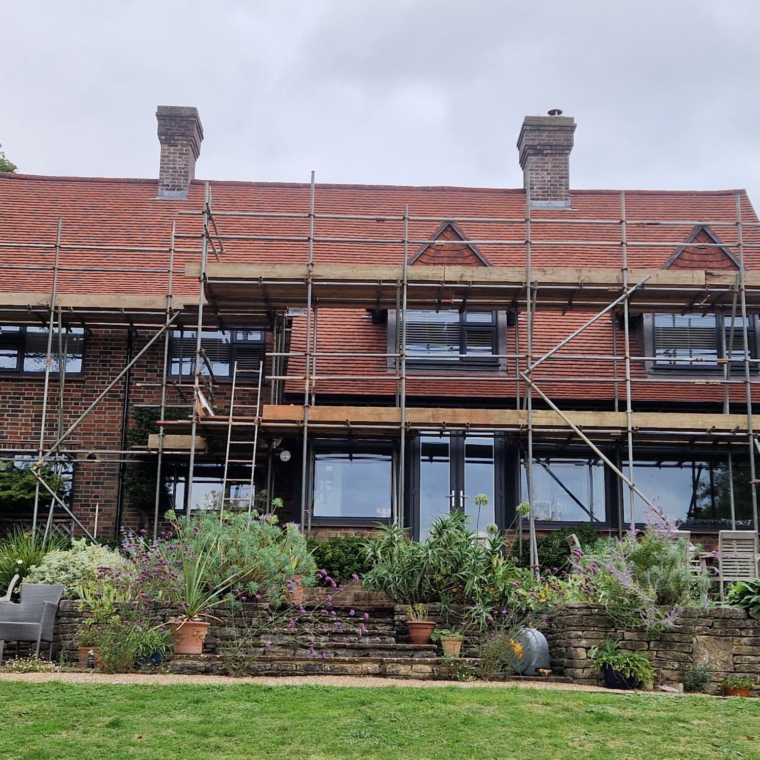 A two-story brick house with a red tiled roof is partially surrounded by scaffolding. The front yard has various green plants and flowers, and a chair is visible on the left side. The sky is cloudy.