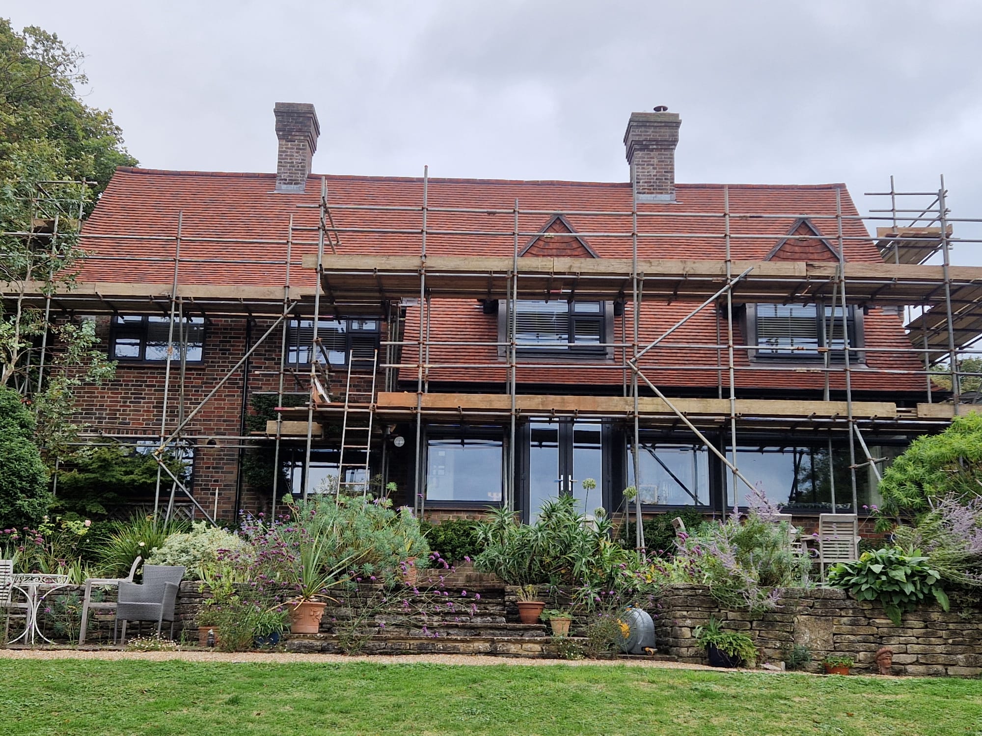 A two-story brick house with a red tile roof is surrounded by scaffolding for renovation. The garden in the foreground has lush greenery and potted plants. The sky above is overcast.