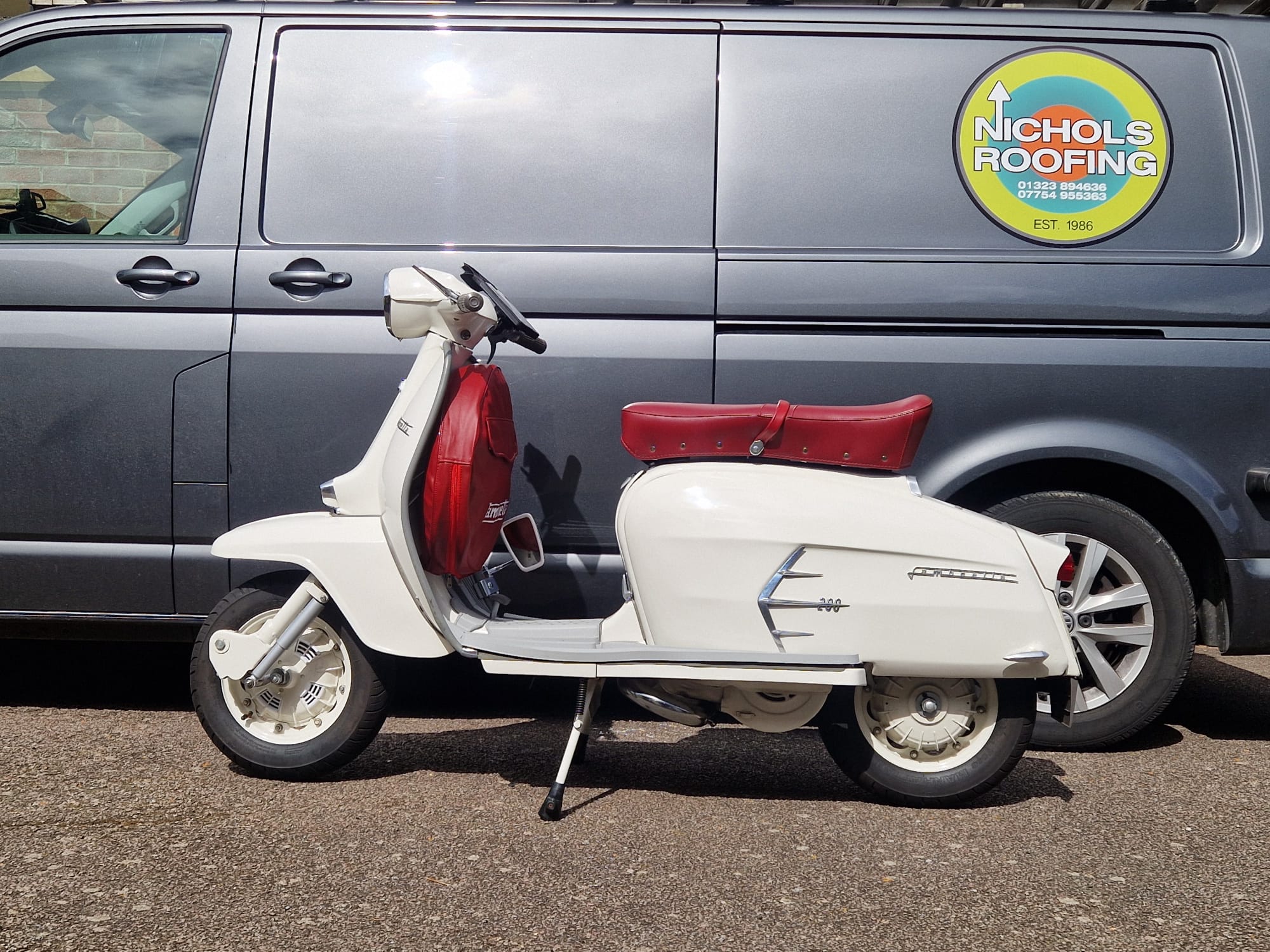 A vintage white scooter with a red seat is parked on the street in front of a dark gray van with "Nichols Roofing" written on a circular logo on the side.