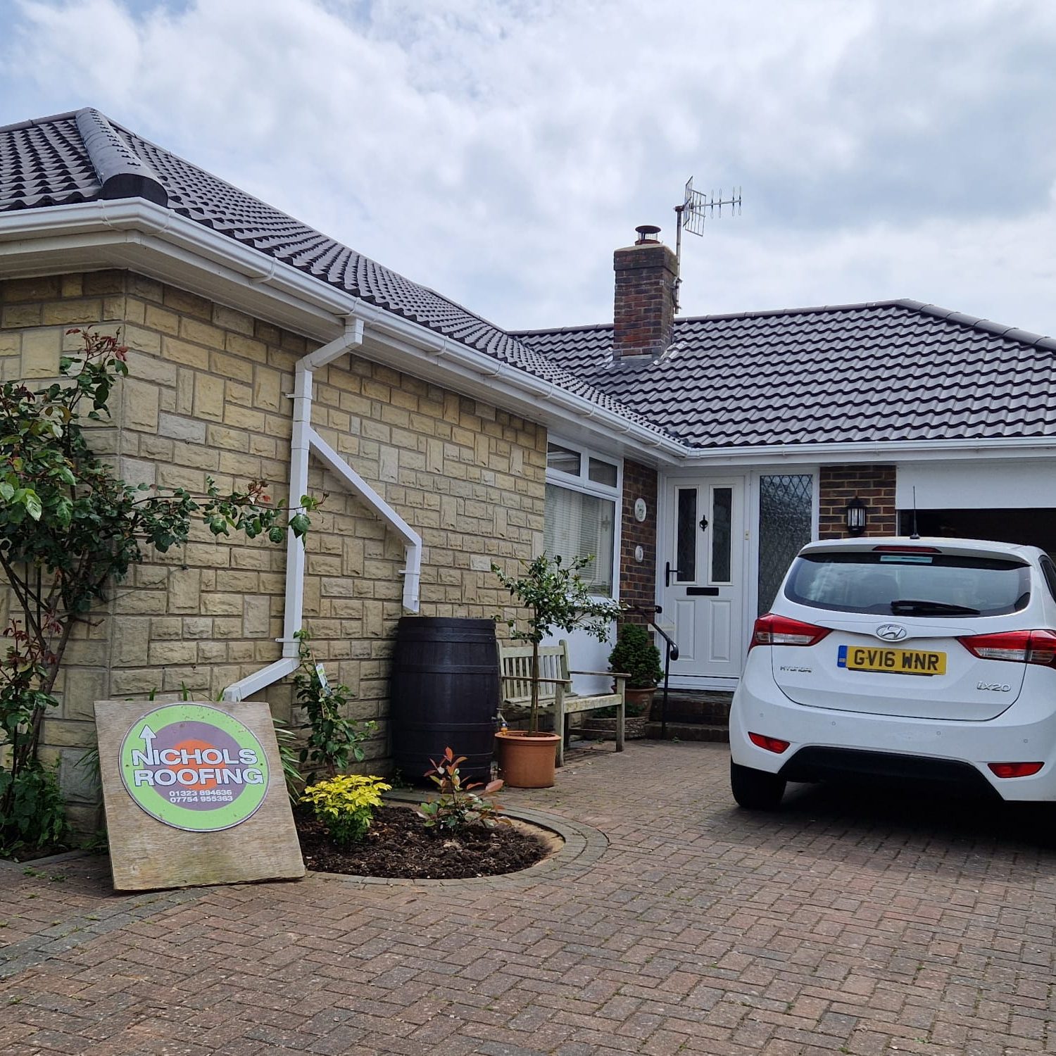 A bungalow with a brick exterior and tiled roof. A white SUV is parked in the paved driveway. A sign for "Nicholls Roofing" is placed on the ground by a small garden with a tree and potted plants near the house entrance.