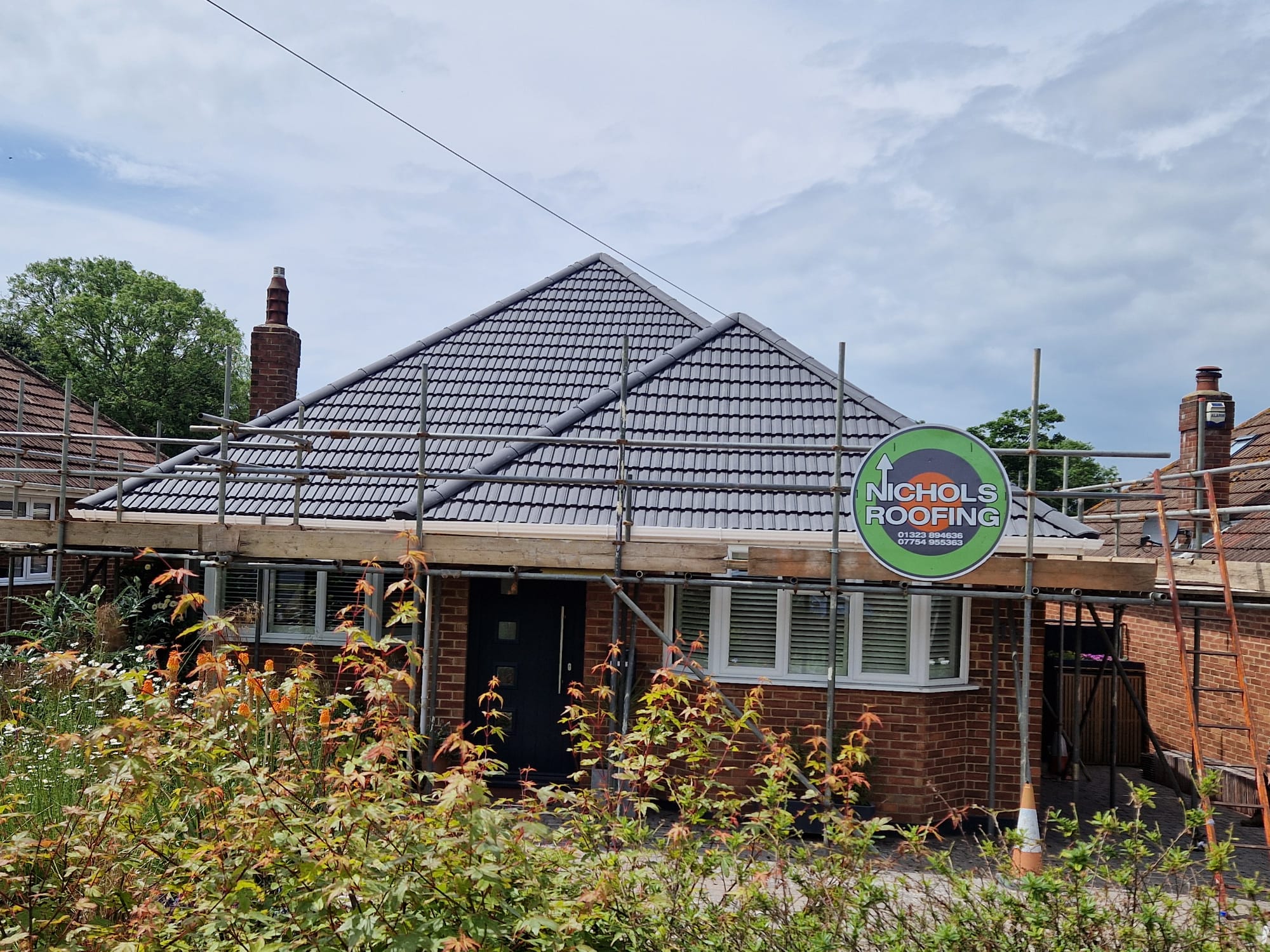 A brick house with a newly tiled roof is surrounded by scaffolding. A sign on the scaffolding reads "Nichols Roofing." Bushes with green and orange foliage are visible in the foreground, and there are trees in the background.