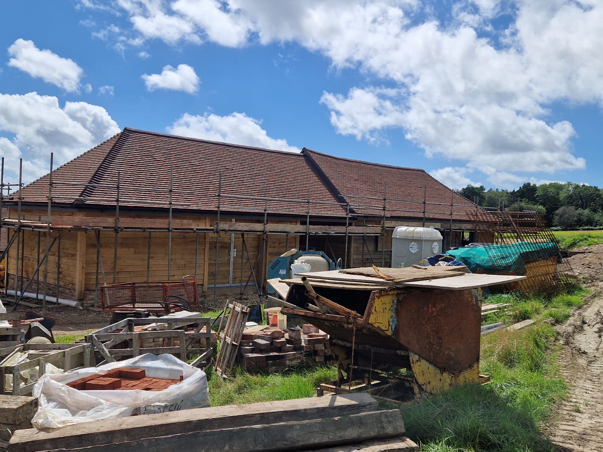 A construction site with a partially built house featuring a tiled roof and wooden walls. Scaffolding surrounds the structure, and construction materials, including bricks and equipment, are scattered in the foreground under a partly cloudy sky.