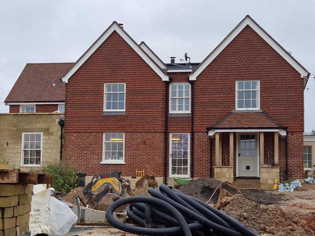 A large two-story brick house under construction, with piles of dirt, building materials, and construction equipment in the foreground. The house features a steep roof and multiple gables, and the weather appears cloudy.