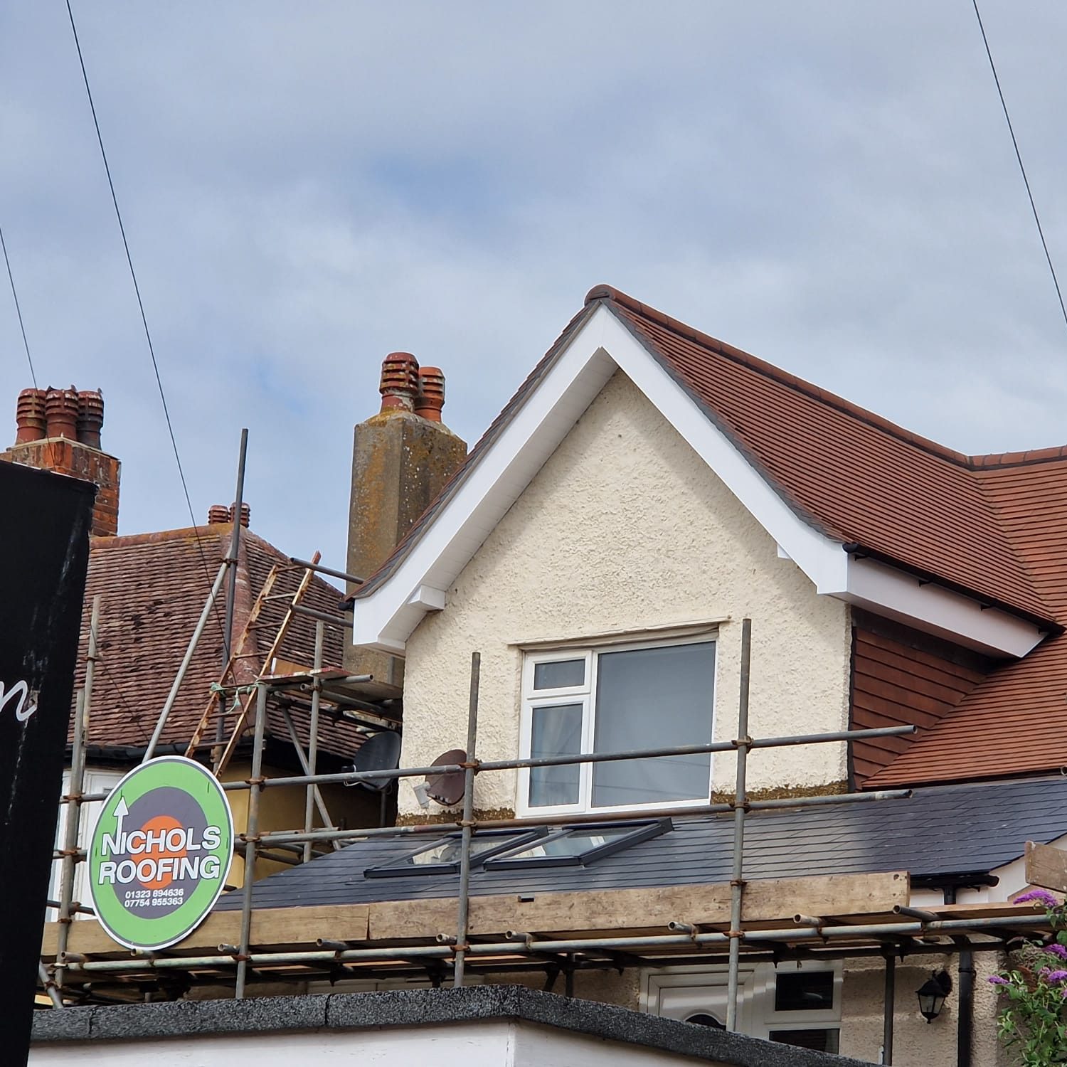 A house with a steep, brown-tiled roof is under repair, with scaffolding set up around it. A sign reads "Nichols Roofing" with a phone number. The sky is overcast, and two brick chimneys are visible.