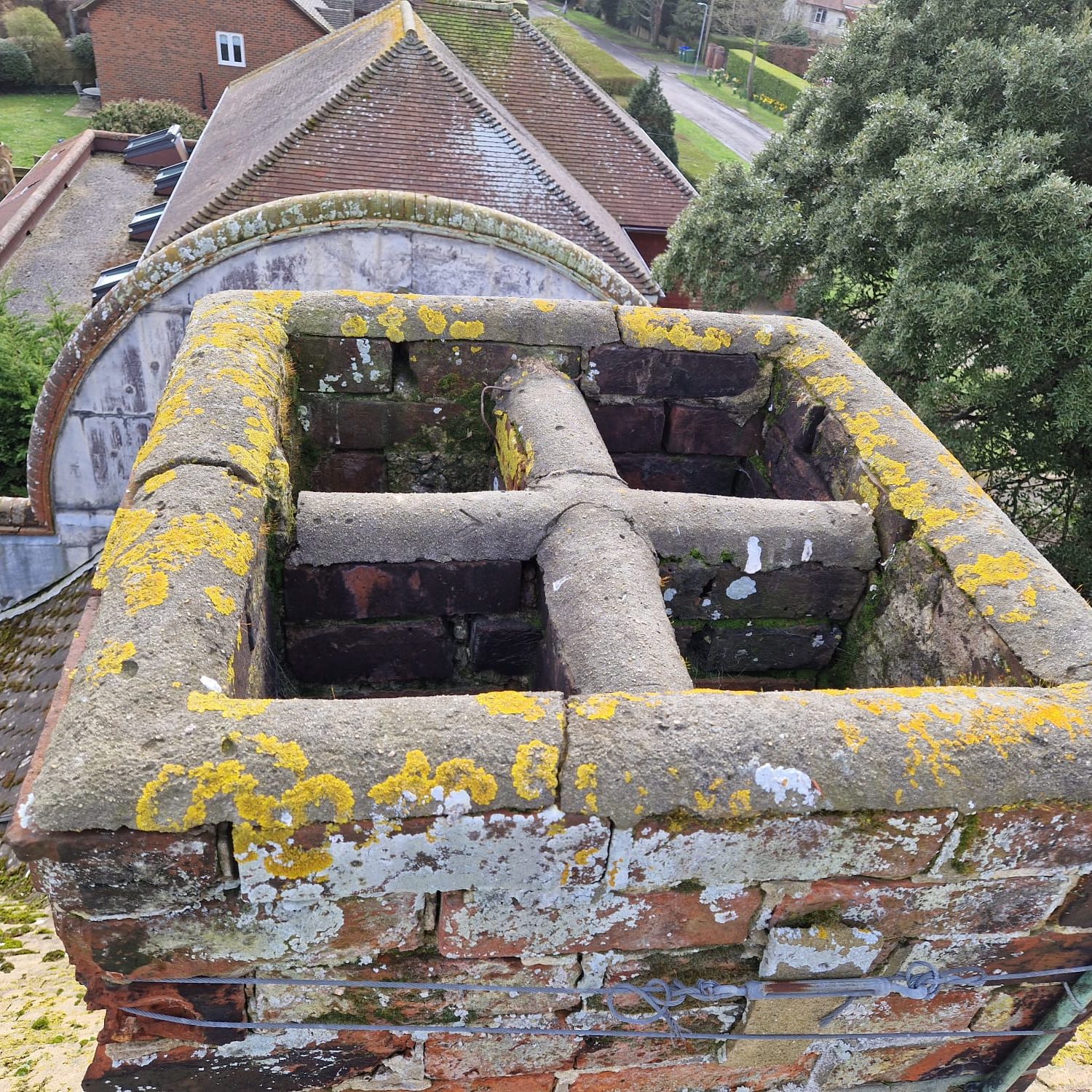 A large, weathered brick chimney with a cross-shaped terracotta flue and patches of yellow lichen on top of a building. The background shows a sloped roof with moss and a tree-lined street.