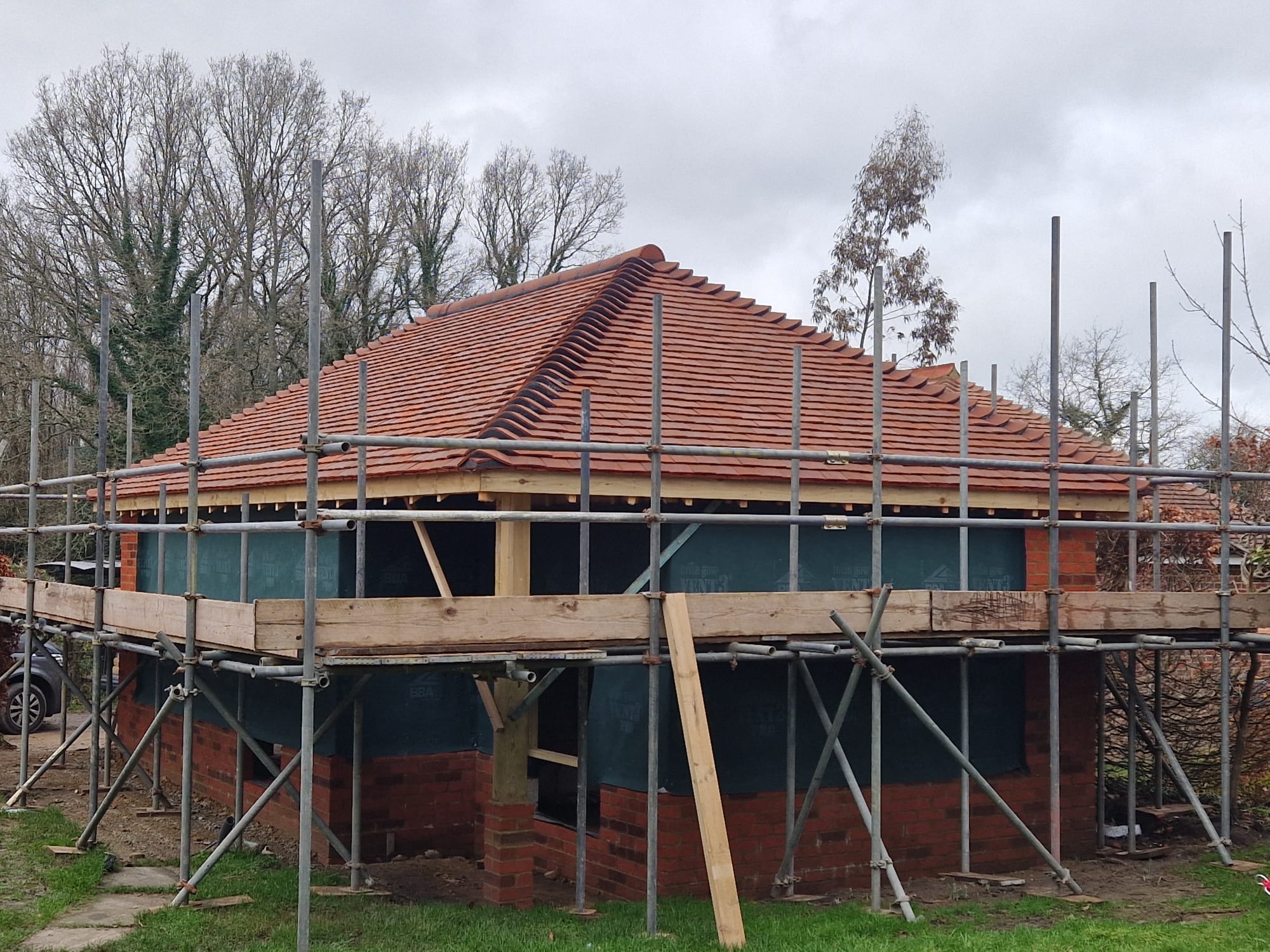 A brick building under construction with a tiled roof partially covered by scaffolding. The structure is surrounded by trees, with a cloudy sky in the background.