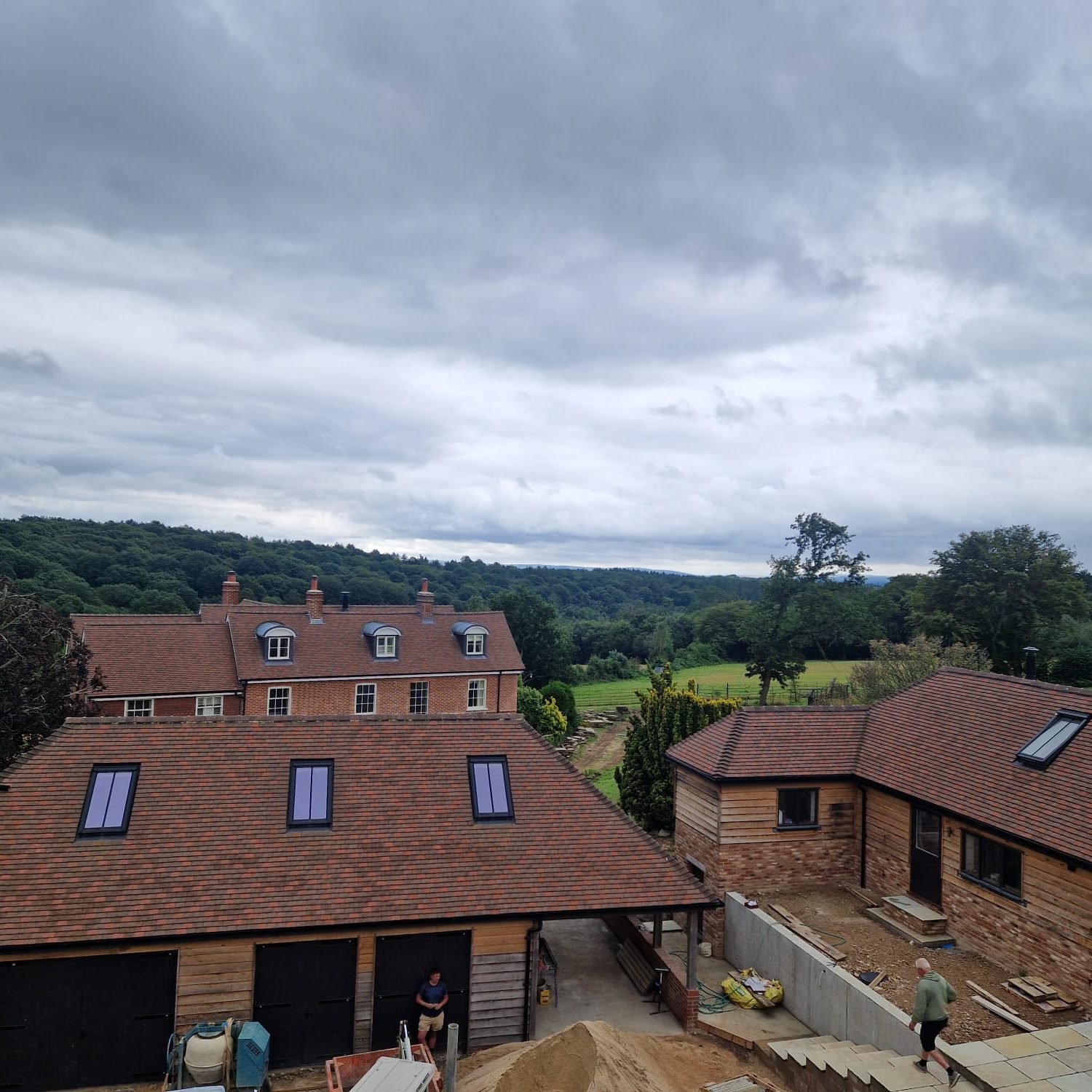 A rural scene with a large brick house and several smaller outbuildings featuring tiled roofs and skylights. Construction materials and a person are visible in the foreground, while lush greenery and a cloudy sky form the backdrop.