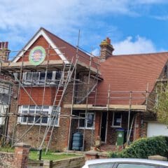 A two-story house with scaffolding surrounding the red-tiled roof for roofing work. A sign on the scaffolding reads "Nichols Roofing." The brick and wood exterior is visible, and trees line the front yard under a clear sky.