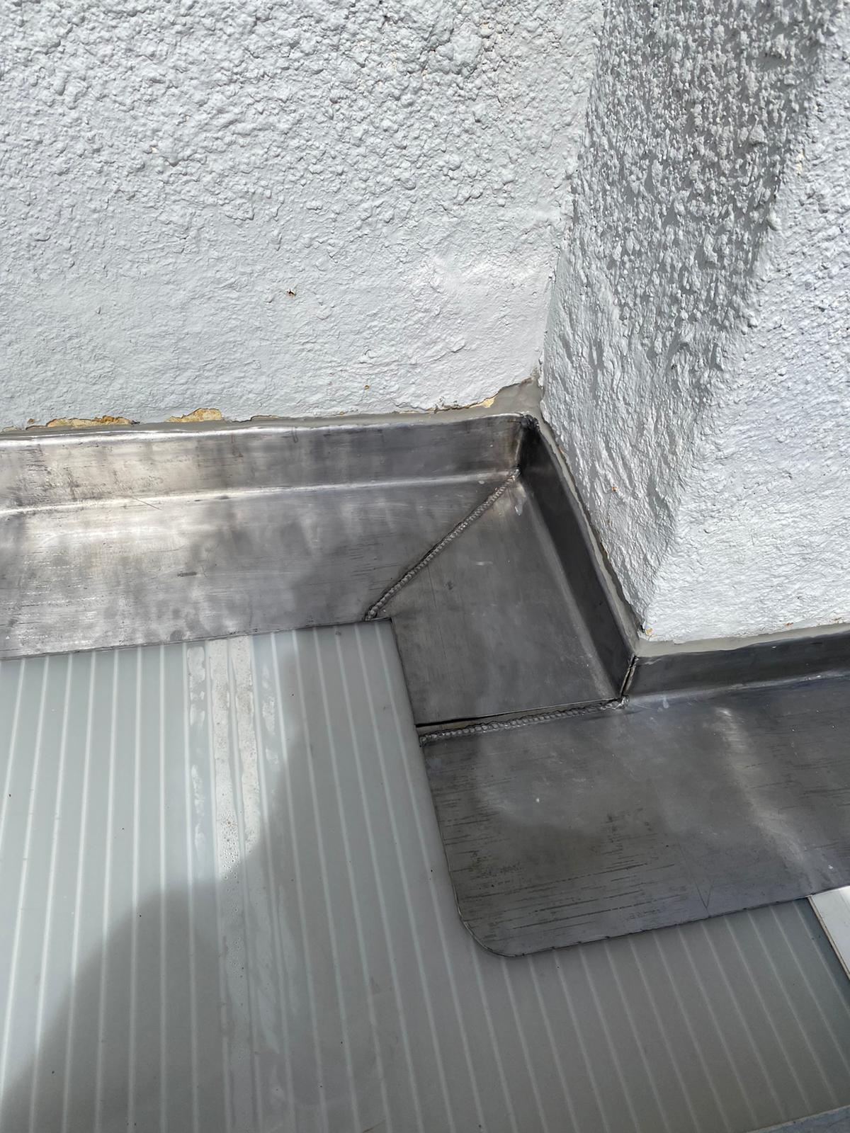 Close-up of a metallic corner flashing on a rooftop or terrace. The flashing is neatly installed where a textured wall meets a slanted surface, likely for waterproofing. The shiny metal contrasts with the light gray wall and floor.