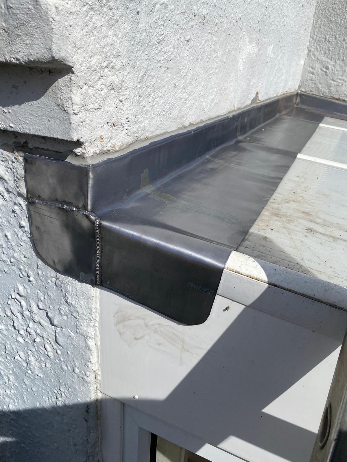 Close-up of a roof corner with a metal flashing installed above a window. The wall is textured and painted light gray. The metal appears shiny and reflective under sunlight. Small patches of wear and discoloration are visible on the metal surface.