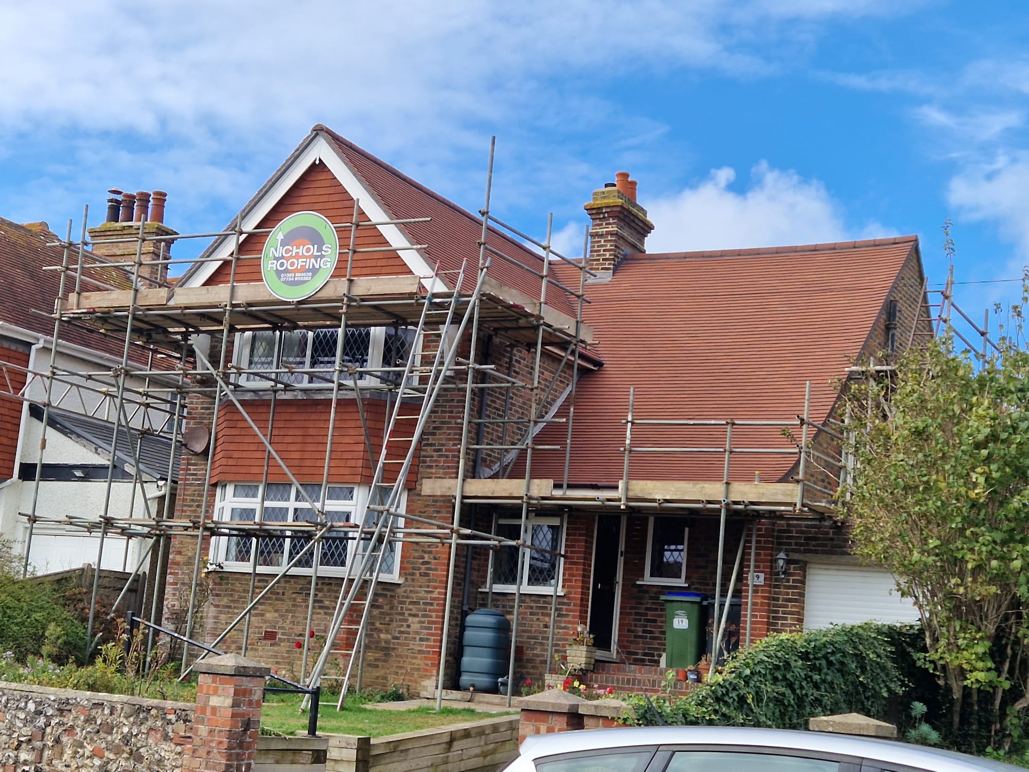 A two-story house with scaffolding surrounding the red-tiled roof for roofing work. A sign on the scaffolding reads "Nichols Roofing." The brick and wood exterior is visible, and trees line the front yard under a clear sky.