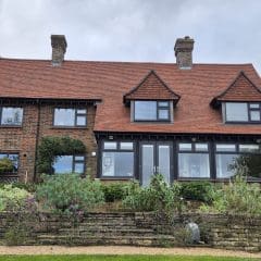 A two-story brick house with a red-tiled roof and three dormer windows. The front features large windows and a variety of plants in a landscaped garden. Two chimneys rise from the roof, and the sky is overcast.