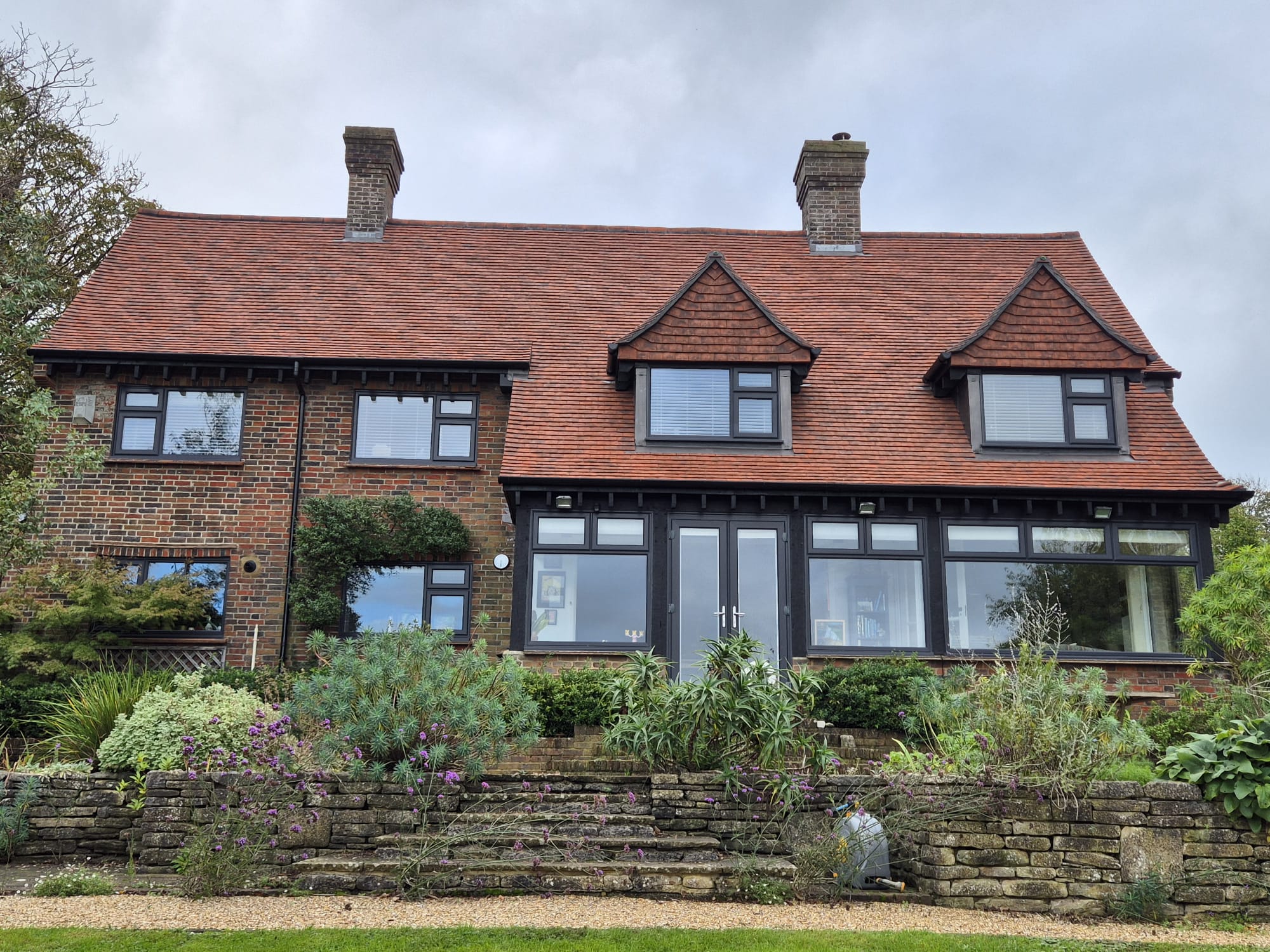 A two-story brick house with a red-tiled roof and three dormer windows. The front features large windows and a variety of plants in a landscaped garden. Two chimneys rise from the roof, and the sky is overcast.