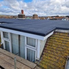 A flat rooftop extension with fresh black roofing is shown. Surrounding houses with sloped roofs are visible in the background under a partly cloudy sky. Wooden scaffolding is present along the building's edge.