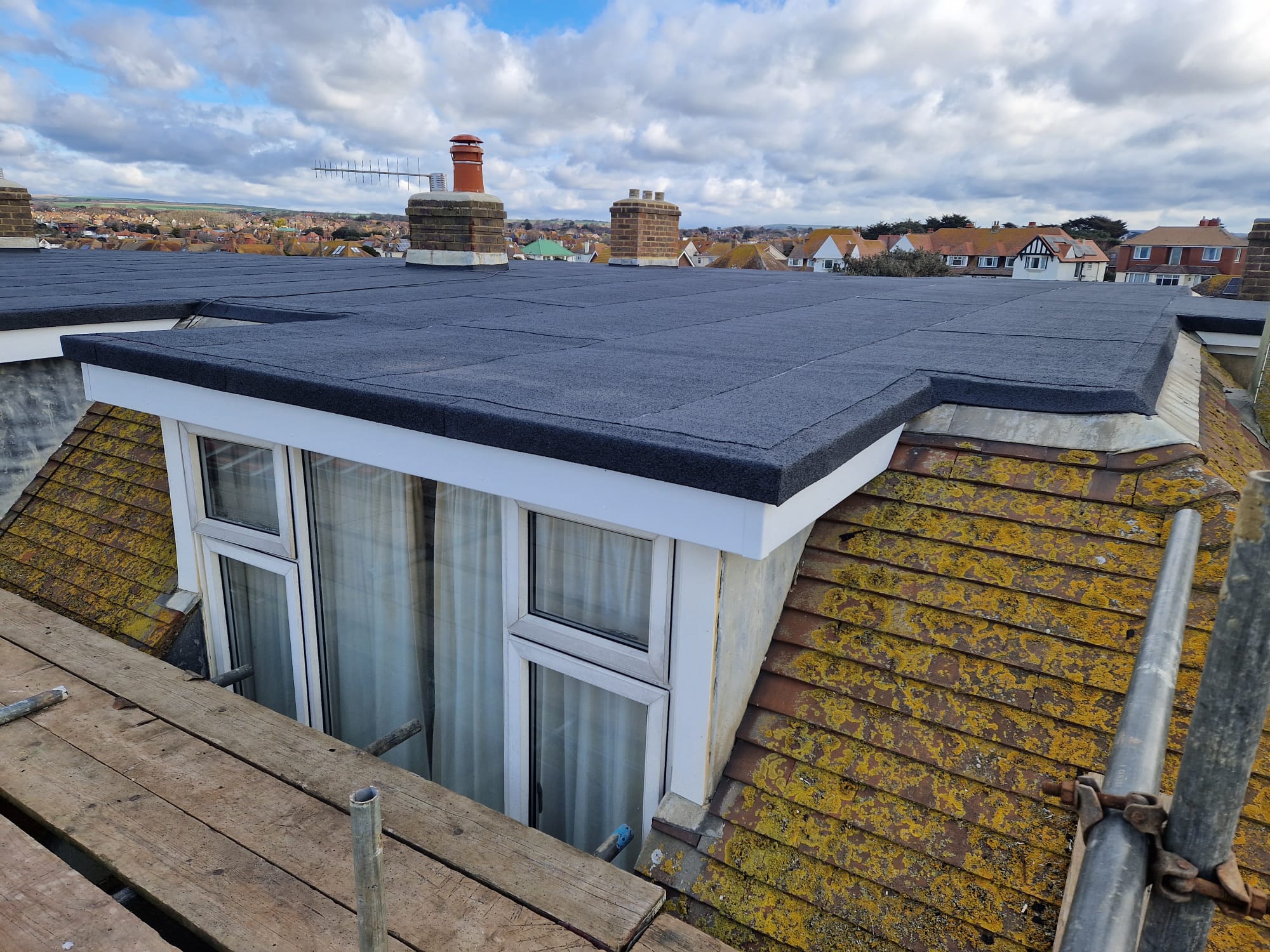 A flat rooftop extension with fresh black roofing is shown. Surrounding houses with sloped roofs are visible in the background under a partly cloudy sky. Wooden scaffolding is present along the building's edge.