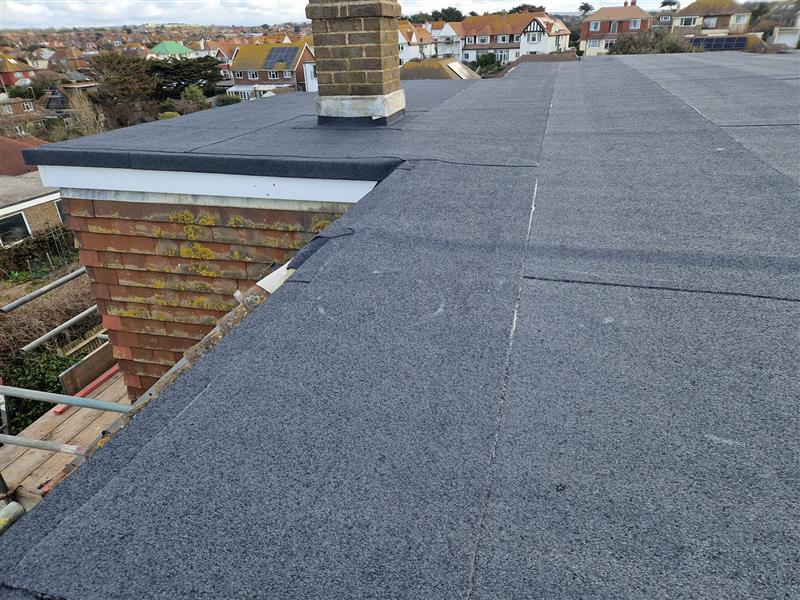 Flat roof covered with dark asphalt shingles, featuring a chimney. Surrounding neighborhood includes rooftops of neighboring houses with a mix of shingles and tiles. Scaffold visible on the left side. Cloudy sky in the background.