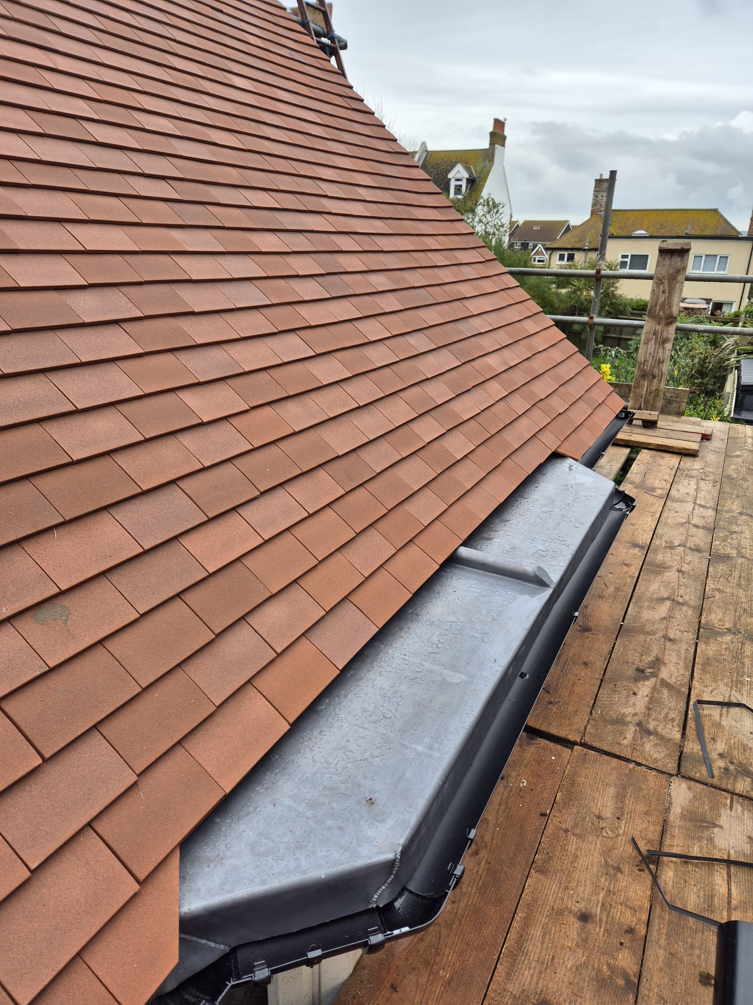 A rooftop with new brown shingles is shown with a scaffolding platform beside it. The sky is overcast, and houses are visible in the background.